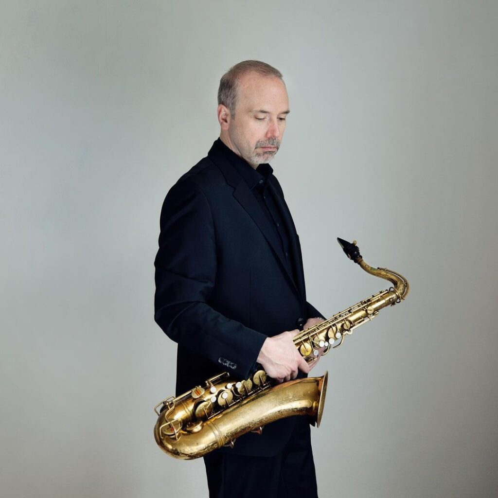 Andreas van Zoelen holding a tenor saxophone. He is looking downwards, wearing a dark suit.